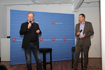 Ministerpräsident und Moderator stehen vor einer blauen Logowand mit Wappen und Schriftzug Niedersachsen