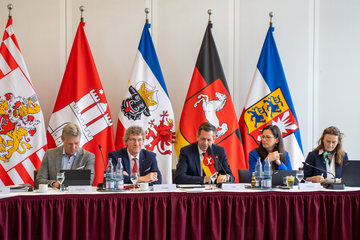 Ministerpräsident Olaf Lies sitzt an einem Tisch und spricht bei der Konferenz der Nordländer. Neben ihm Vertreterinnen der Niedersächsischen Staatskanzlei und im Hintergrund sind die Flaggen der Nordländer aufgereiht.