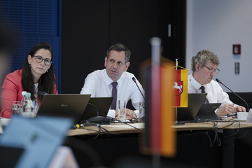 Ministerpräsident Olaf Lies sitzt an einem Tisch und spricht zu den Anwesenden der Ministerpräsidentenkonferenz. Im Vordergrund sind schemenhaft eine kleine Flagge von Deutschland und etwas deutlicher eine Niedersachsenflagge zu erkennen.
