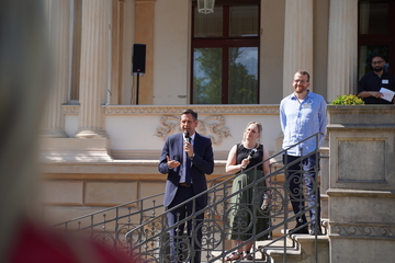 Ministerpräsident Olaf Lies steht auf eine Treppe mit geschwungenem Geländer und spricht zu Gästen, neben ihm eine Frau mit einem Mikrofon und ein Mann, beide in sommerlicher Kleidung.