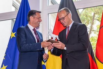 Ministerpräsident Olaf Lies überreicht Bundeskanzler Friedrich Merz eine kleine weiße Pferdestatue. Im Hintergrund sind die Flaggen der EU, Deutschlands und Niedersachsens aufgereiht.