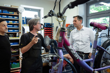 Ministerpräsident Olaf Lies steht neben zwei Monteuren einer Fahrradwerkstatt. Im Vordergrund ist ein Kinderfahrrad zu sehen, im Hintergrund Aufbewahrungsmöglichkeiten für Werkzeug und Zubehör.