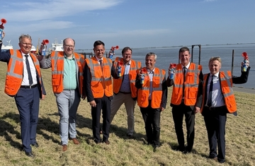Ministerpräsident Olaf Lies und Umweltminister Christian Meyer stehen mit Vertretern von NPorts mit Warnwesten am Strand. Im Hintergrund sind entfernt Schiffe und Wasser zu sehen.
