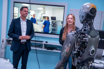 Ministerpräsident Lies schaut auf ein Exoskelett, während eine Mitarbeiterin die Funktion erklärt. Im Hintergrund ist ein Sichtfenster, durch das ein Krankenhausbett zu sehen ist, an dem zwei Pflegekräfte stehen.