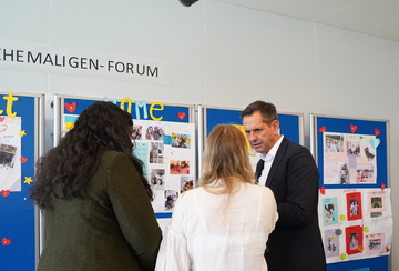 Ministerpräsident Lies steht vor einer Wandzeitung, auf der Kinder und Jugendliche Bilder und Geschichten aus ihrer Heimat Mykolajiw angepinnt haben. Eine Jugendliche steht mit dem Rücken zur Kamera und erzählt von ihrem Leben.