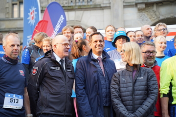 Ministerpräsident Liest steht neben einem Polizisten und einer Frau und lächelt. Im Hintergrund sind verschiedene Läuferinnen und Läufer zu sehen, die auf den Start des Laufes der Rettungskräfte Run4Respect warten.