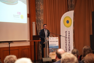 Ministerpräsident Lies hält eine Rede an einem Pult, rechts von ihm ist ein Aufsteller und links eine Leinwand