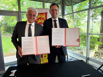 Der Präsident der Region Normandie, Hervé Morin, und Ministerpräsident Olaf Lies. stehen in einem lichtdurchfluteten Raum mit großen Fenstern und halten jeweils aufgeschlagene, unterzeichnete Dokumente in den Händen.