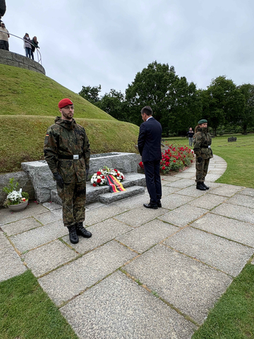 Ministerpräsident Olaf Lies steht in dunklem Anzug vor einem steinernen Gedenkbereich mit niedergelegtem Kranz in Schwarz-Rot-Gold. Zwei uniformierte Soldaten flankieren die Szene im Stillstand.