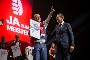 Ministerpräsident Lies steht mit einem Meisterabsolventen auf der Bühne. Der Meister hält seinen Meisterbrief in der Hand und streckt seinen Arm in die Luft.