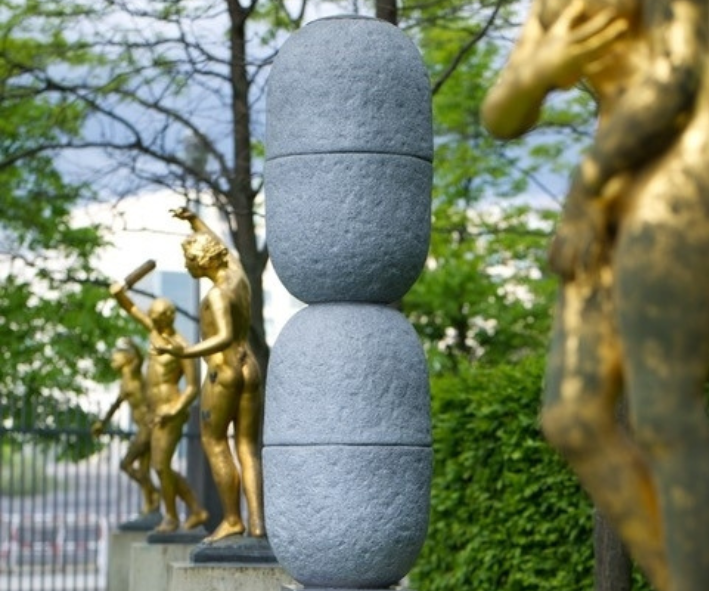 Das Bild zeigt eine moderne, graue Skulptur zwischen goldenen Barockstatuen. Sie besteht aus zwei abgerundeten zylindrischen Formen, die aufeinandergesetzt wurden.