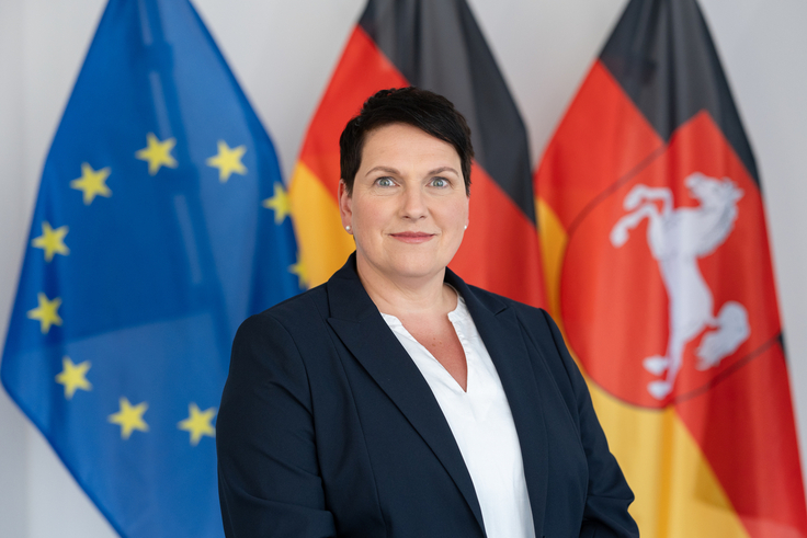 Portraitaufnahme von Ministerin Melanie Walter. Im Hintergrund sind schemenhaft die Europa-, Deutschland- und Niedersachsenflagge zu sehen.