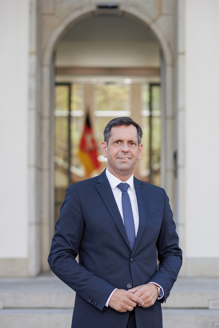 Ministerpräsident Olaf Lies steht vor der Niedersächsischen Staatskanzlei. Im Hintergrund zeichnet sich schemenhaft die Niedersachsenflagge ab.