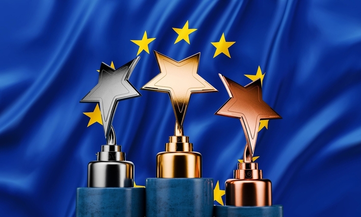 Drei Pokale mit farblich entsprechenden Sternen in Gold, Silber und Bronze stehen auf einzelnen Podesten. Im Hintergrund ist die Europaflagge eingeblendet.