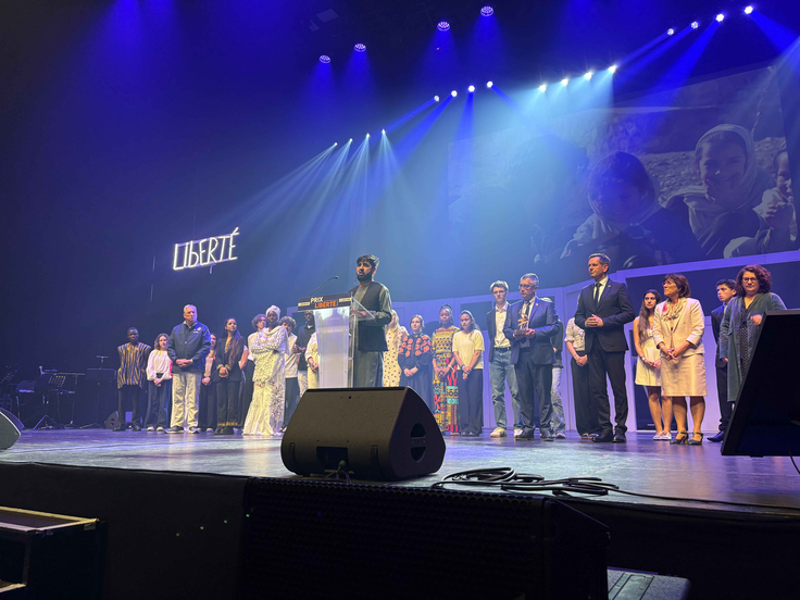 Bühne mit blauem Bühnenlicht, eine Person steht am Rednerpult mit der Aufschrift „Prix Liberté“, dahinter Menschen in einer Reihe. Rechts davon stehen offiziell gekleidete Personen. Im Hintergrund ist groß das Wort „LIBERTÉ“ beleuchtet.