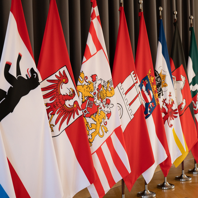 Flaggen der Bundesländer stehen aufgereiht vor einem dunklen Vorhang.