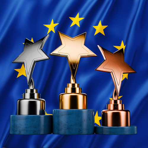 Drei Pokale mit farblich entsprechenden Sternen in Gold, Silber und Bronze stehen auf einzelnen Podesten. Im Hintergrund ist die Europaflagge eingeblendet.