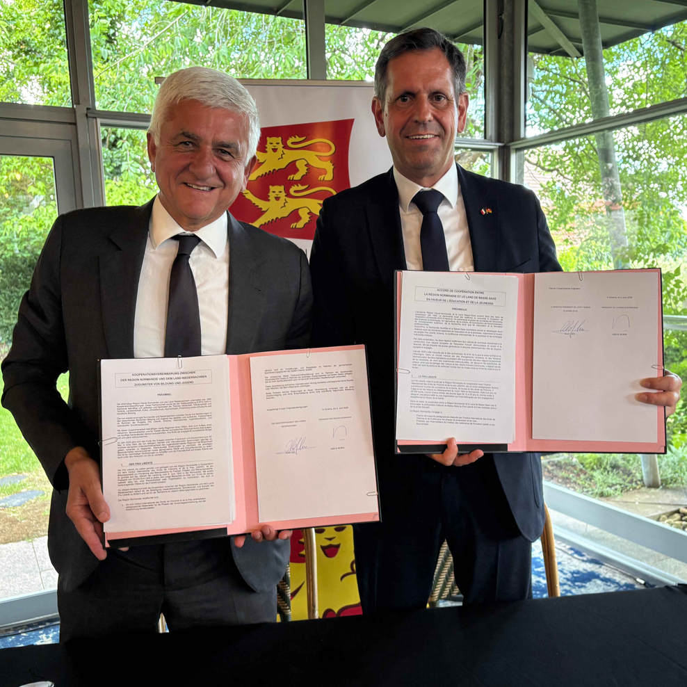 Der Präsident der Region Normandie, Hervé Morin, und Ministerpräsident Olaf Lies. stehen in einem lichtdurchfluteten Raum mit großen Fenstern und halten jeweils aufgeschlagene, unterzeichnete Dokumente in den Händen.