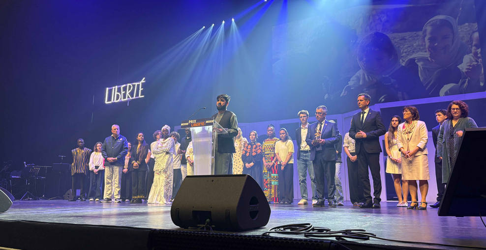 Bühne mit blauem Bühnenlicht, eine Person steht am Rednerpult mit der Aufschrift „Prix Liberté“, dahinter Menschen in einer Reihe. Rechts davon stehen offiziell gekleidete Personen. Im Hintergrund ist groß das Wort „LIBERTÉ“ beleuchtet.